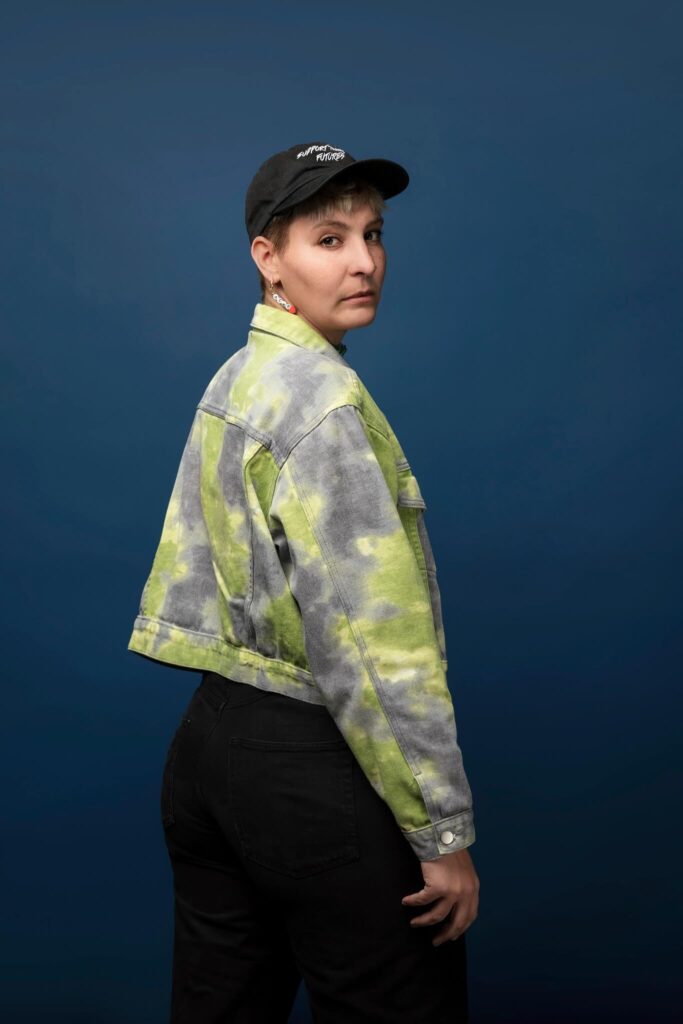 lovislovislovis, original copy, dj, a white nonbinary person with a green washed jacket and a black hat saying support trans futures, looking over their shoulder into the camera with a serious gaze